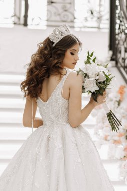 Gelinlik giymiş, açık omuzlu, uzun kahverengi saçlı, elinde bir buket çiçekle bir gelin suretinde bir kız.