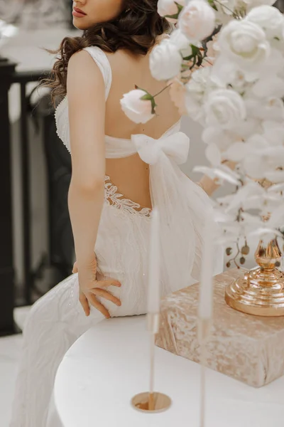 漂亮的新娘拿着一个花瓶坐在桌旁 这个女人穿着一件有花边和敞着背的婚纱 背上绑着一个蝴蝶结 — 图库照片