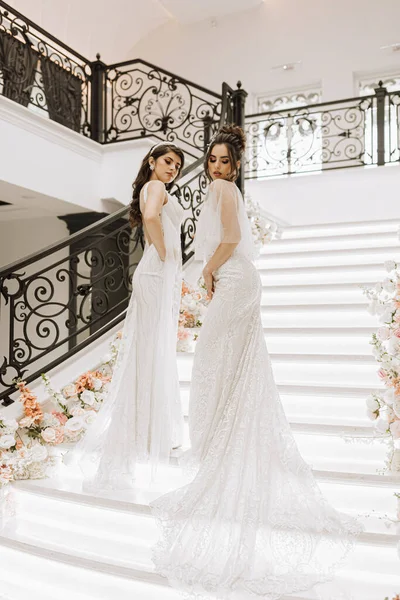 Two beautiful girls in wedding dresses pose on the stairs of a restaurant decorated with flowers. Photo shoot for a wedding salon