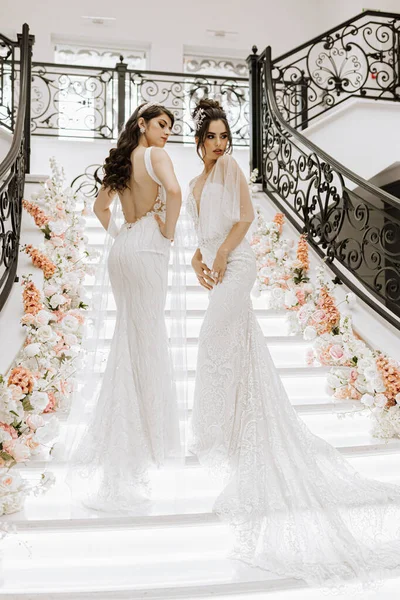 Two beautiful girls in wedding dresses pose on the stairs of a restaurant decorated with flowers. Photo shoot for a wedding salon