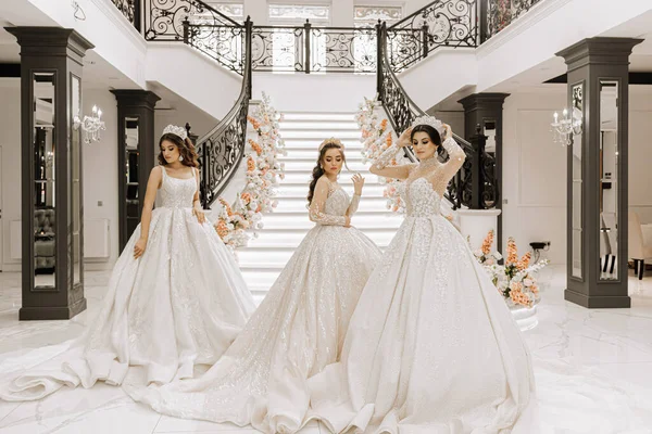 Three beautiful girls in luxurious wedding dresses with long trains pose in the hall near the steps of a restaurant decorated with flowers. Photo session for a wedding salon. Stylish fashion