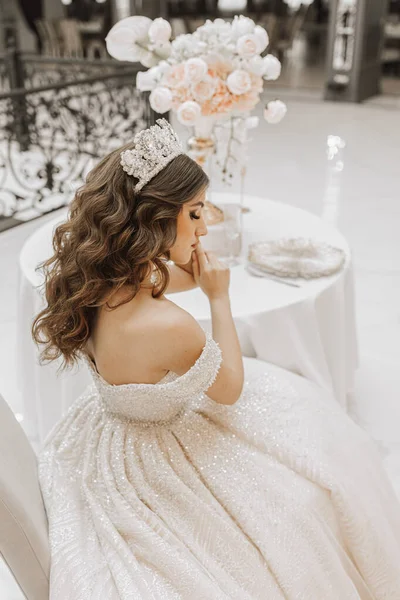studio photo of a princess bride from the back in a wedding dress with luxurious hair and makeup, the bride is sitting at the table