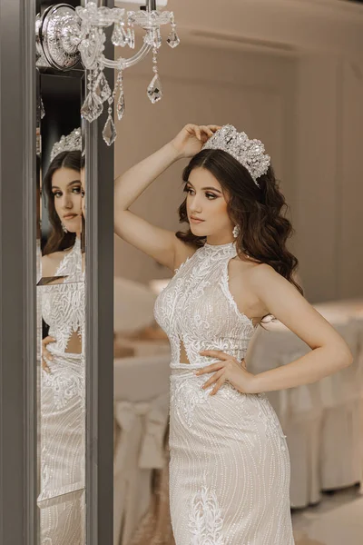 Portrait of a girl in a wedding dress near a mirror, with a crown on her head. The concept of a royal feast. Vertical photo.