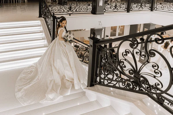 一个身穿婚纱 手持长长的火车和一束玫瑰的女孩在楼梯上的迷人的摄影棚照片 华丽的内部 广角照片 — 图库照片