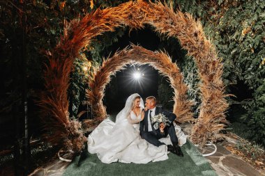 Gelin ve damadın portresi. Gelin ve damat mısır kulaklarından yapılmış bir kemerin arka planında oturuyorlar. Elbisenin uzun treni. Gönüllü duvak. Kristal mücevherler. Kırsal biçim