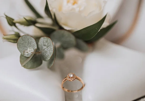 A gold wedding ring with a diamond near a rose with greenery, on a white background. Wedding details in the photo.