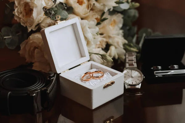 Wedding details on a brown background. Men\'s watch, cufflinks and black belt. Wedding bouquet, wedding rings in a white box decorated with flowers.
