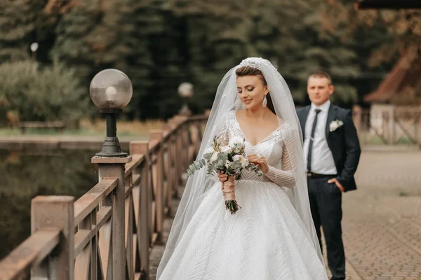 結婚式の肖像画 長いドレスのブルネットの花嫁は 彼女の花束を保持しています ピーニーと緑 古典的なスーツのグロムは 橋の彼女の後ろを歩きます — ストック写真