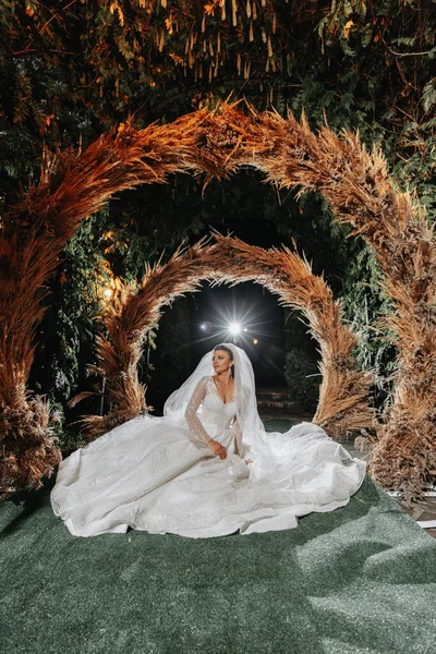 Portrait of the bride. The bride is sitting against the background of an arch made of ears of corn. Long train of the dress. Voluminous veil. Crystal jewelry. Rustic style