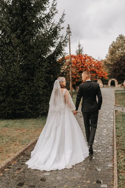 秋の公園で恋をするカップル ブロンドの花嫁が袖と花束を手にしたウェディングドレス グロムはクラシックなブラックスーツ ホワイトシャツ ネクタイです リアビュー — ストック写真
