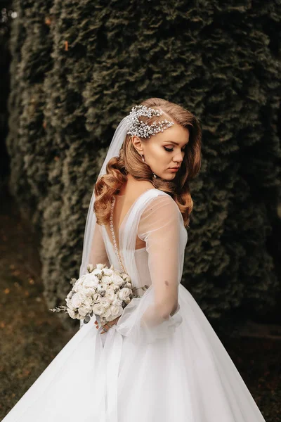スリーブ付きのウェディングドレスの花嫁の写真 美しい髪型と彼女の頭にティアラ 彼女の背後にある花束を保持 — ストック写真