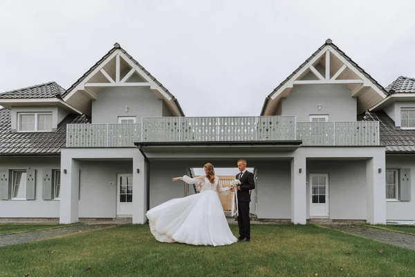 a couple in love on the background of gray houses. A blonde bride in a wedding dress with sleeves throws up her dress. Groom in a classic black suit, white shirt and tie. Smiles and joy