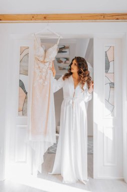 Gelin düğün için hazırlanıyor. Uzun siyah saçlı bir kız gelinliğinin yanında duruyor.