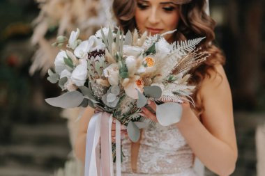 Gelinin portresi. Gelin çiçeklerden ve kurumuş çiçeklerden yapılmış bir kemerin arkasında duruyor. Şık bir gelinlik. Düğün süslemeleri. Kırsal biçim