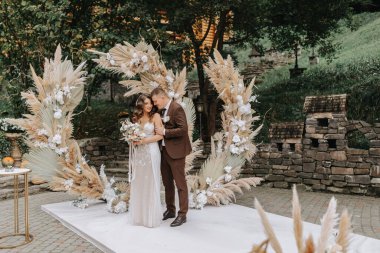 Gelin ve damat eğleniyor. Çiçeklerle süslenmiş bir kemerin altında duran düğün buketiyle yeni evliler ve dışarıda kurumuş çiçekler..