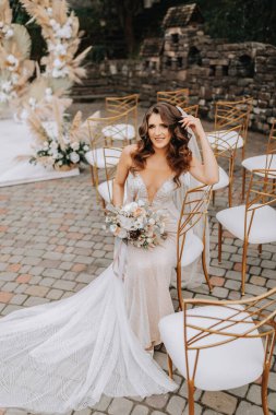 Elinde bir buket çiçekle gelin, düğün töreninden sonra beyaz sandalyelerde oturur ve altın kenarlı pozlar verir.