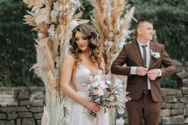 Portrait of the bride and groom. The bride stands against the background of the groom and an arch made of flowers and dried flowers. Stylish wedding dress. Wedding decorations. Rural style