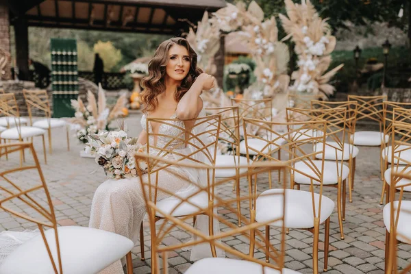 Bride with a bouquet of flowers sits and poses on white chairs with gold edges after the wedding ceremony