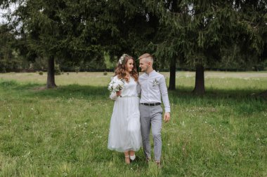 Ormanda düğün yürüyüşü. Gelin ve damat yeşil çimlerin üzerinde yürür ve birbirlerine bakarlar. Geniş açı fotoğrafı.