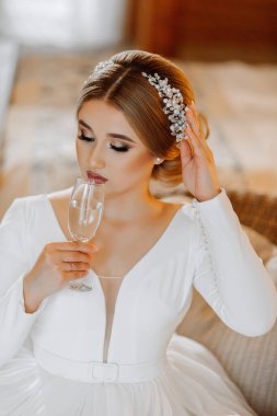 Bir otel odasında elinde bir bardak şampanyayla beyaz gelinlik içinde güzel bir gelinin portresi.