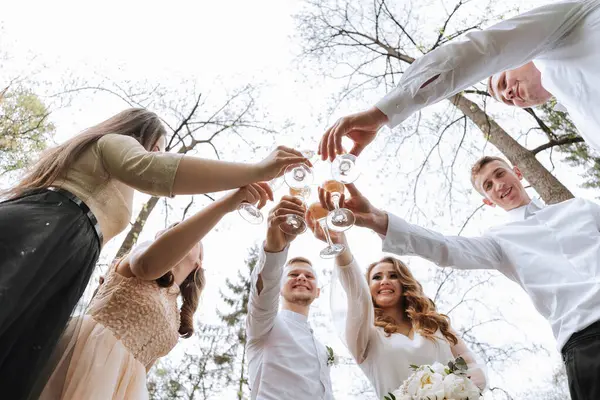 The bride and groom and their friends pose against the background of the forest. A large group of people are having fun at their friends' wedding. They drink champagne from glasses