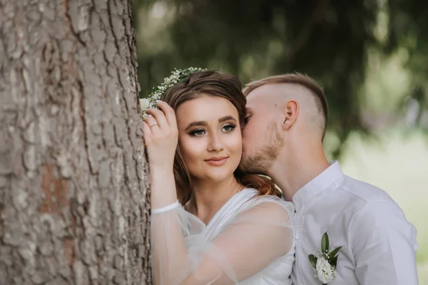 Portrait of the bride and groom. A bride with long hair and a wreath on her head near a tall tree, from behind the groom hugs her and kisses her on the cheek.