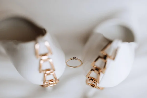 An elegant gold wedding ring with a diamond, standing between women\'s white high-heeled shoes. Photo from above on a white background
