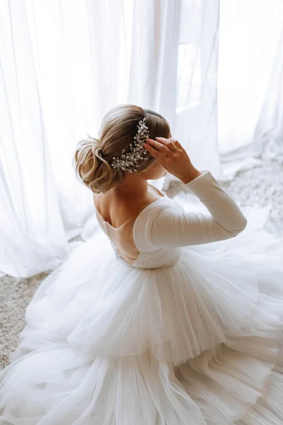 Portrait of a beautiful bride in a white wedding dress with a long train in a hotel room. The bride is sitting on the floor against a large window. Photo of the model from behind