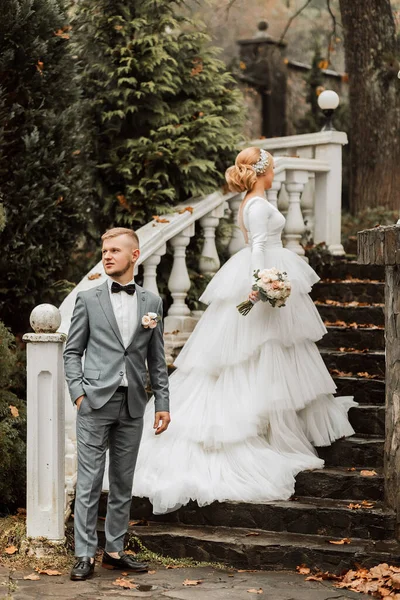 Portrait of the bride and groom. The bride in a wedding dress with a train on the elegant steps. Groom in a classic gray suit, white shirt and bow tie. Full length photo. Wedding in a top location