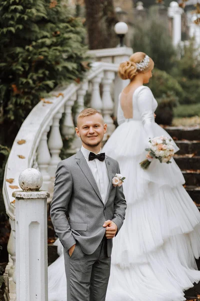 Portrait of the bride and groom. The bride in a wedding dress with a train on an elegant staircase. Groom in a classic gray suit, white shirt and bow tie. Full length photo. Wedding in a top location