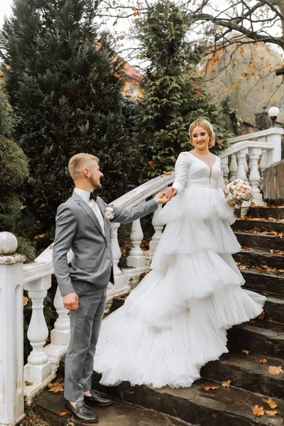 Portrait of the bride and groom. The bride in a wedding dress with a train on an elegant staircase. Groom in a classic gray suit, white shirt and bow tie. He holds her hand. Wedding in a top location