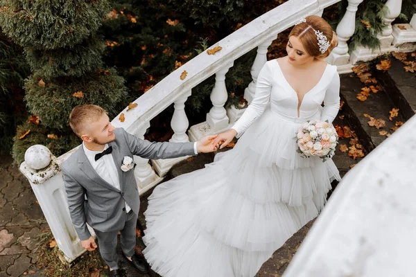 Portrait of the bride and groom. The bride in a wedding dress with a train on an elegant staircase. Groom in a classic gray suit, white shirt and bow tie. He holds her hand. Wedding in a top location
