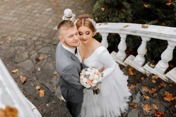 Portrait of the bride and groom from above. The bride in a wedding dress on an elegant staircase. Groom in a classic gray suit, white shirt and bow tie. He takes it away. Wedding in a top location