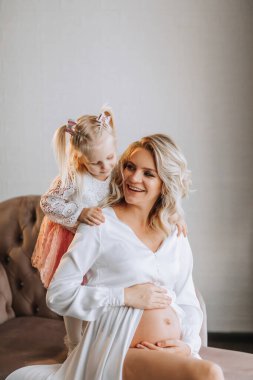 Beyaz elbiseli güzel hamile bir kadının stüdyo portresi. Küçük kızıyla birlikte koltukta oturuyor.