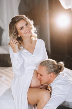 Beyaz elbiseli, trenle yatakta oturan, yanında hamile karnını öpen güzel hamile bir kadının stüdyo portresi.