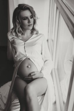Stüdyo portresi, yakın plan, güzel hamile bir kadının beyaz bir yatakta, hamile karnını elleriyle tutarken.
