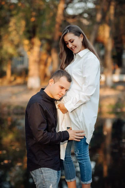 穿着白衬衫的年轻孕妇 丈夫把手放在妻子的肚子上 靠在耳朵上 — 图库照片