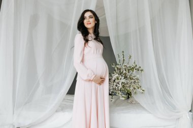 Pembe elbiseli güzel hamile bir kadın ellerini karnında tutuyor. Sevgi dolu bir anne bir çocuğun doğmasını bekliyor. Hamilelik, annelik, hazırlık ve bekleme kavramı.