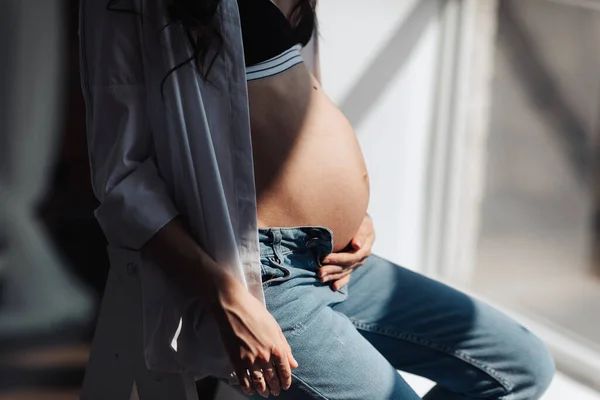 modern beautiful pregnant woman in jeans and shirt hugs her tummy with hands in beautiful sunlight. Concept of pregnancy, motherhood, preparation and waiting. The beauty of a woman during pregnancy