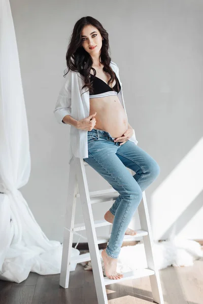 modern beautiful pregnant woman in jeans and shirt hugs her tummy with hands in beautiful sunlight. Concept of pregnancy, motherhood, preparation and waiting. The beauty of a woman during pregnancy