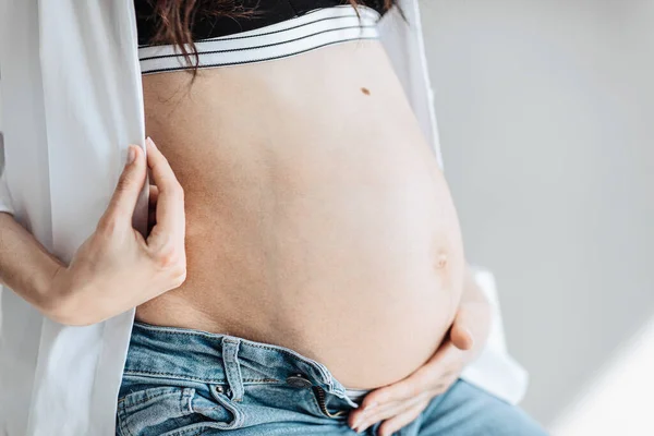 modern beautiful pregnant woman in jeans and shirt hugs her tummy with hands in natural light. Concept of pregnancy, motherhood, preparation and waiting. The beauty of a woman during pregnancy