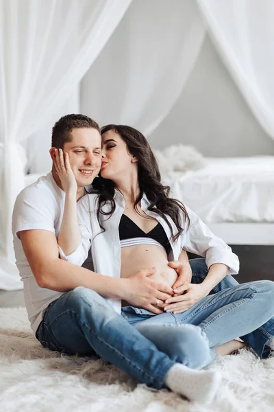 modern beautiful pregnant woman in jeans and shirt with her husband at home, caring, hugging, kissing. Concept of pregnancy, motherhood, preparation and waiting. The beauty of a woman during pregnancy