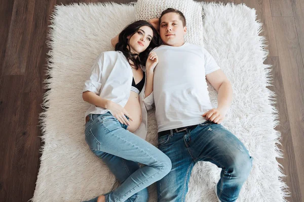modern beautiful pregnant woman in jeans and shirt with her husband on the bed. Photo from above. Concept of pregnancy, motherhood, preparation and waiting. The beauty of a woman during pregnancy