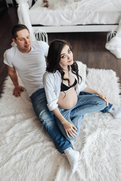 modern beautiful pregnant woman in jeans and shirt with her husband at home, caring, hugging, kissing. Concept of pregnancy, motherhood, preparation and waiting. The beauty of a woman during pregnancy