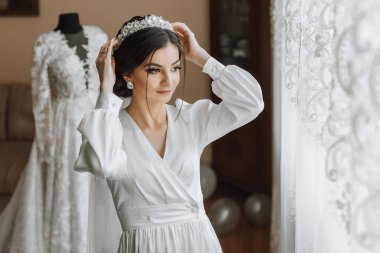 Kafasında taç olan bornozlu güzel bir kızın portresi. Moda, cazibe, kavramlar. Gelinin sabahında, bornozlu gelin gelinliğiyle bir mankenin yanında duruyor.