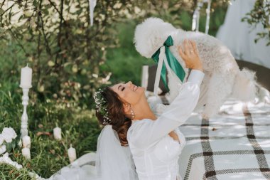 Beyaz elbiseli genç, zarif bir gelin kafasında taze çiçeklerle bahçede fino köpeğiyle poz veriyor.