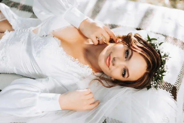 木の下の庭のベッドに横たわっている夢のような花嫁の肖像画 花嫁は白いドレスと彼女の頭に新鮮な花のティアラを着ています — ストック写真