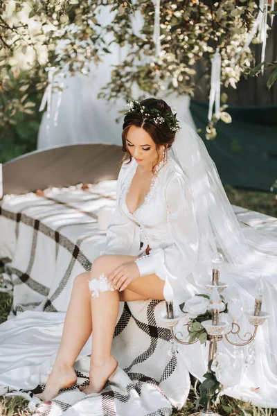 新娘躺在花园里的床上的早晨 新娘穿着一件白色的衣服 头上戴着一束鲜花的头冠 腿上戴着网状的吊袜带 — 图库照片