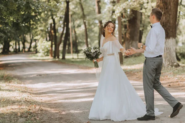 一对年轻夫妇新婚夫妇正在一个夏季公园散步 新娘头戴花环 身穿漂亮的白色婚纱 新郎头戴白衬衫 — 图库照片