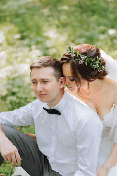 ドレスの美しいかわいい花嫁は緑の草の上に座っている白いシャツの後ろにスタイリッシュなグロムを抱きしめます 結婚式の肖像画 恋の新婚旅行の写真 — ストック写真